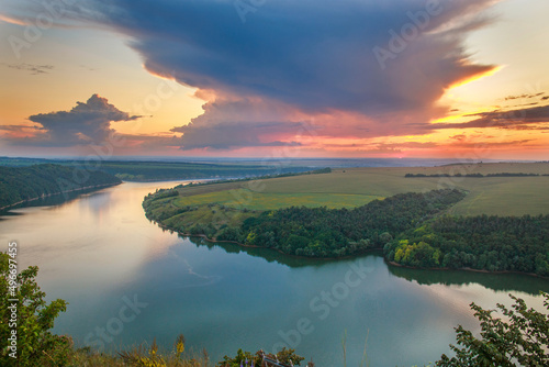 Aerial view river in middle of green forests and fields on background of an epic sunset sky and colorful clouds
