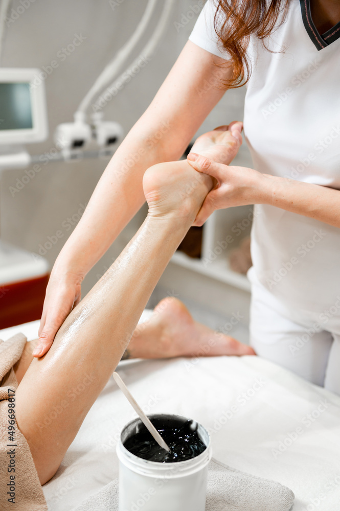 Obraz premium The girl is given an anti-cellulite foot massage with a black scrub cream. Depilation of legs in a beauty salon. Close-up of the massage. Depilation close-up.