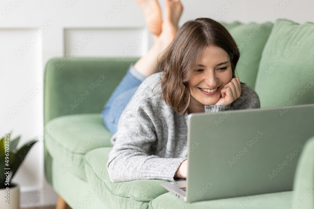 Young beautiful smiling woman using laptop lying on the couch. Concept of freelance job, home office. Female using technology for learning, shopping, e-commerce, communication, social media Young beautiful smiling woman using laptop lying on the couch. Concept of freelance job, home office. Female using technology for learning, shopping, e-commerce, communication, social media