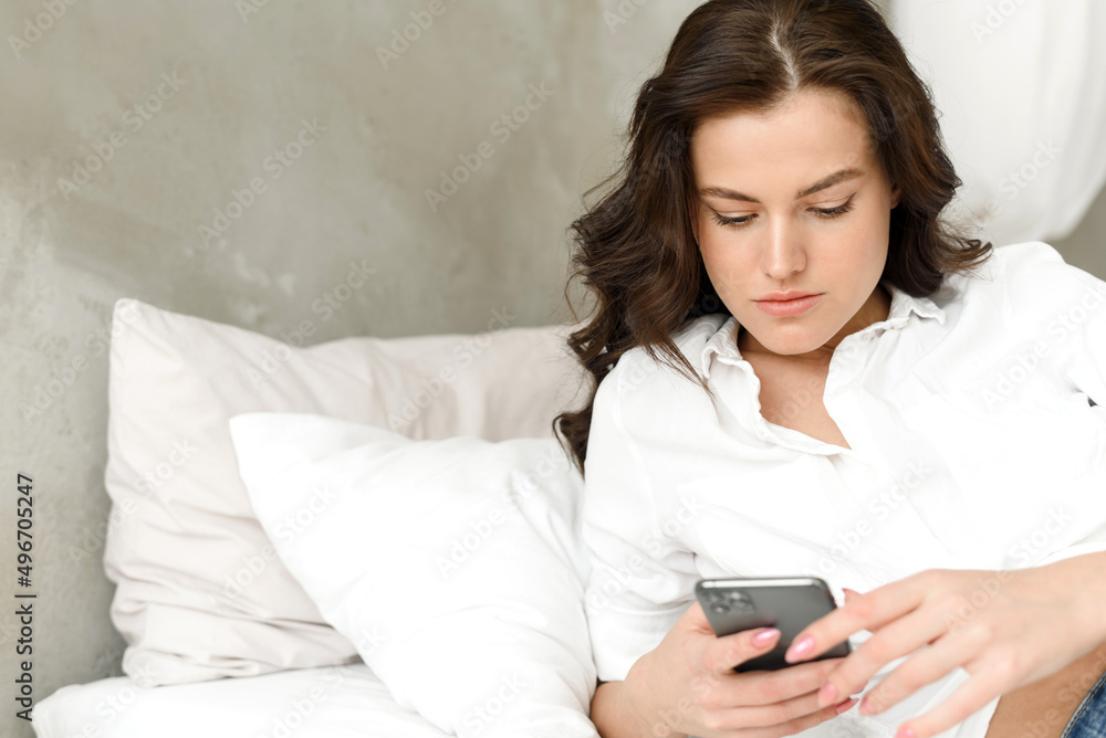 Happy young woman with mobile phone lying on the sofa. Young happy woman lies down on bed and browses social networks at home on smartphone. Happy young woman with mobile phone lying on the sofa. Young happy woman lies down on bed and browses social networks at home on smartphone.