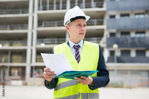Caucasian man engineer in workwear and white hardhat standing on building site and analysing documentation.