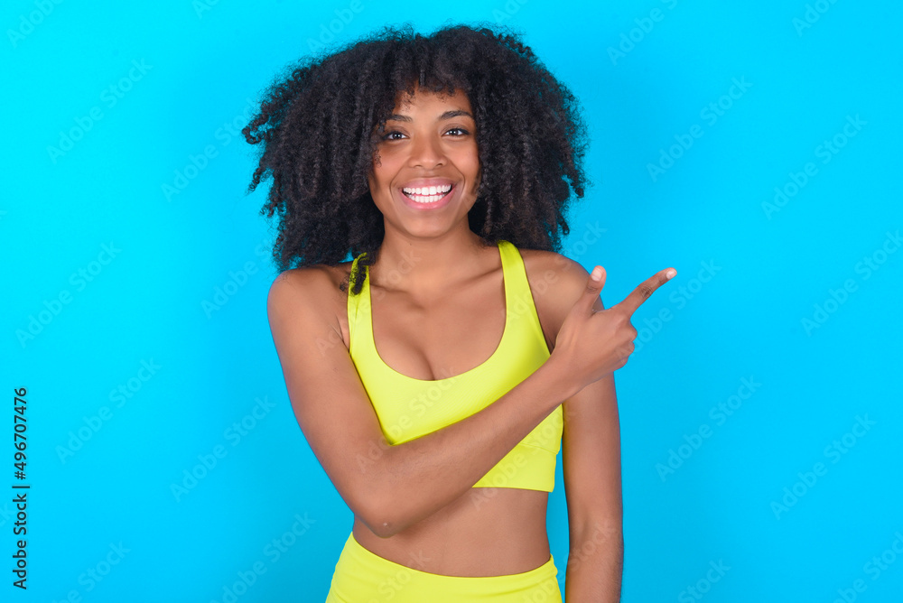 young woman with afro hairstyle in sportswear against blue background pointing away and smiling to you. Look over there! young woman with afro hairstyle in sportswear against blue background pointing away and smiling to you. Look over there!