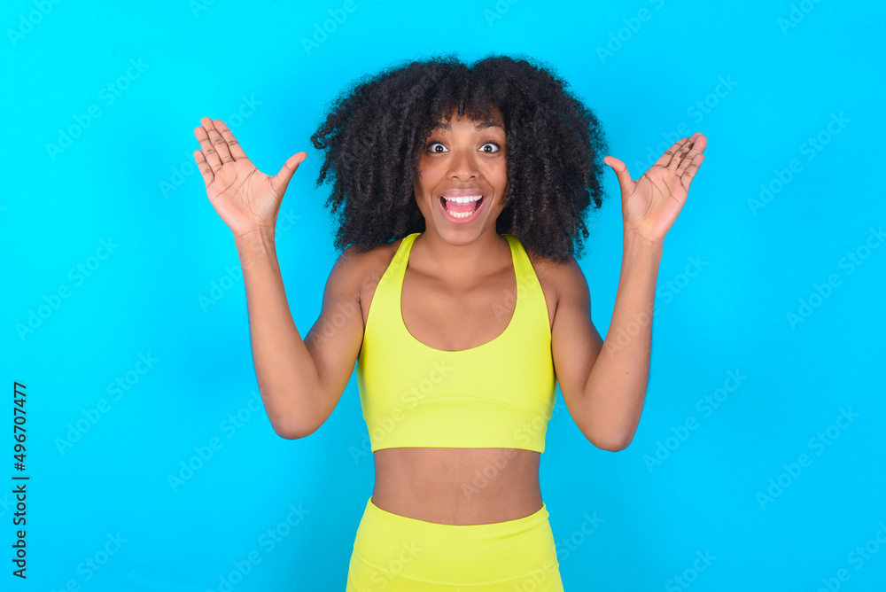 young woman with afro hairstyle in sportswear against blue background raising hands up, having eyes full of happiness rejoicing his great achievements. Achievement, success concept. young woman with afro hairstyle in sportswear against blue background raising hands up, having eyes full of happiness rejoicing his great achievements. Achievement, success concept.