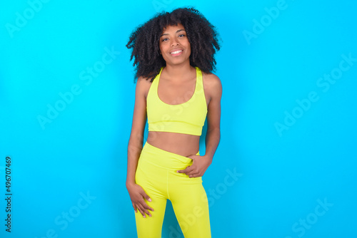 Portrait of successful young woman with afro hairstyle in sportswear against blue background, smiling broadly with self-assured expression. Confidence and business concept.