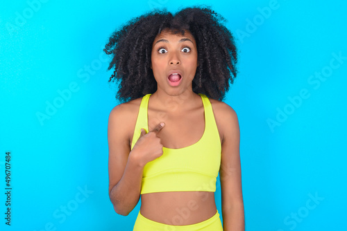 young woman with afro hairstyle in sportswear against blue background being in stupor shocked, has astonished expression pointing at oneself with finger saying: Who me?