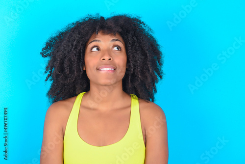 young woman with afro hairstyle in sportswear against blue background with thoughtful expression, looks away keeps hands down bitting his lip thinks about something pleasant.