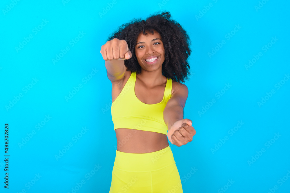 Portrait of charming young woman with afro hairstyle in sportswear against blue background, smiling broadly while holding hands over her head. Confidence and relax concept. Portrait of charming young woman with afro hairstyle in sportswear against blue background, smiling broadly while holding hands over her head. Confidence and relax concept.