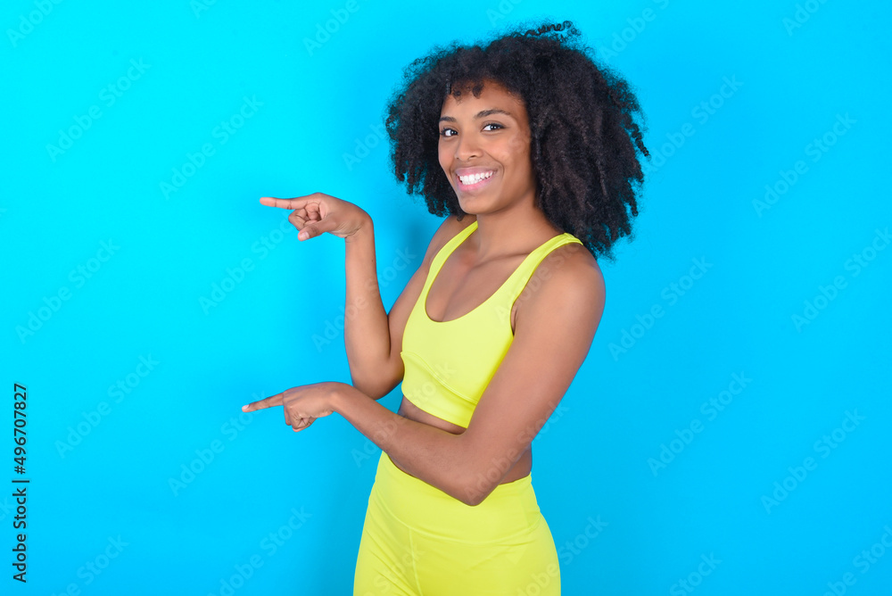 young woman with afro hairstyle in sportswear against blue background points aside with surprised expression with mouth opened, shows something amazing. Advertisement concept. young woman with afro hairstyle in sportswear against blue background points aside with surprised expression with mouth opened, shows something amazing. Advertisement concept.