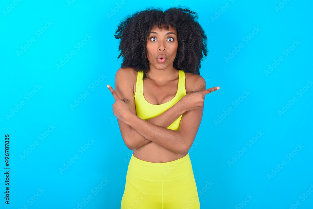Confused young woman with afro hairstyle in sportswear against blue background chooses between two ways, points at both sides with crossed hands, feels doubt. Need your advice. Confused young woman with afro hairstyle in sportswear against blue background chooses between two ways, points at both sides with crossed hands, feels doubt. Need your advice.