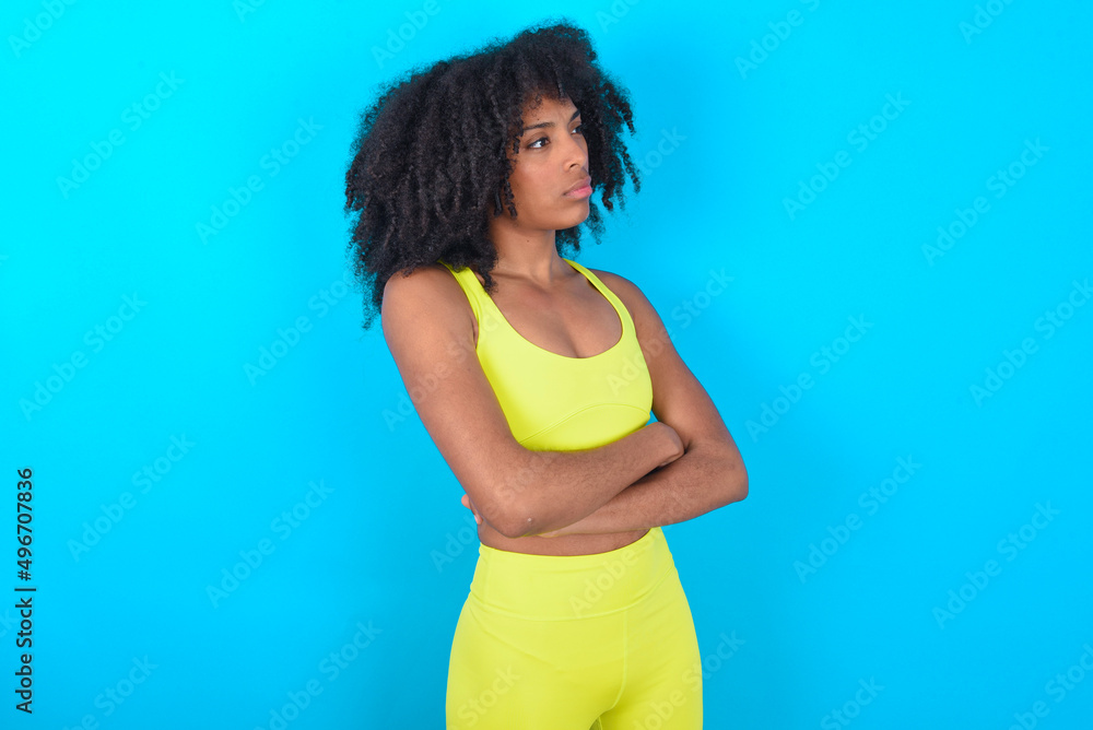 Image of upset young woman with afro hairstyle in sportswear against blue background with arms crossed. Looking with disappointed expression aside after listening to bad news. Image of upset young woman with afro hairstyle in sportswear against blue background with arms crossed. Looking with disappointed expression aside after listening to bad news.