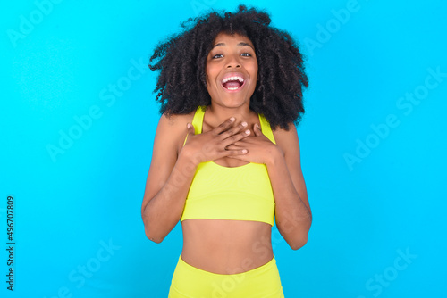 Happy smiling young woman with afro hairstyle in sportswear against blue background has hands on chest near heart. Human emotions, real feelings and facial expression concept.