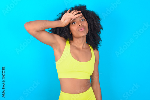 young woman with afro hairstyle in sportswear against blue background wiping forehead with hand making phew gesture, expressing relief feels happy that he prevented huge disaster. It was close enough