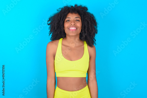 young woman with afro hairstyle in sportswear against blue background with happy and funny face smiling and showing tongue. © Jihan