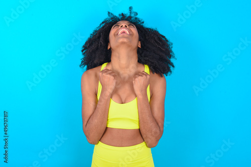 young woman with afro hairstyle in sportswear against blue background looks with excitement up, keeps hands raised, notices something unexpected.