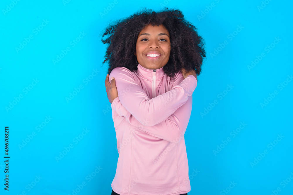 Fototapeta premium Charming pleased young woman with afro hairstyle in technical sports shirt against blue background embraces own body, pleasantly feels comfortable poses. Tenderness and self esteem concept