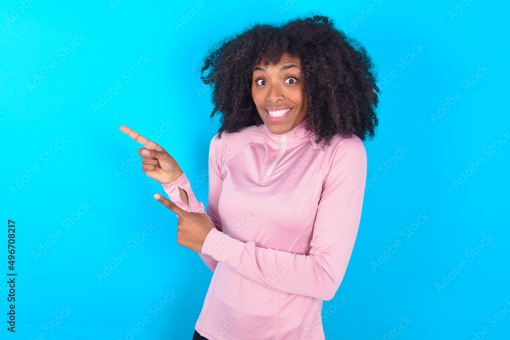 young woman with afro hairstyle in technical sports shirt against blue background points at copy space indicates for advertising gives right direction young woman with afro hairstyle in technical sports shirt against blue background points at copy space indicates for advertising gives right direction