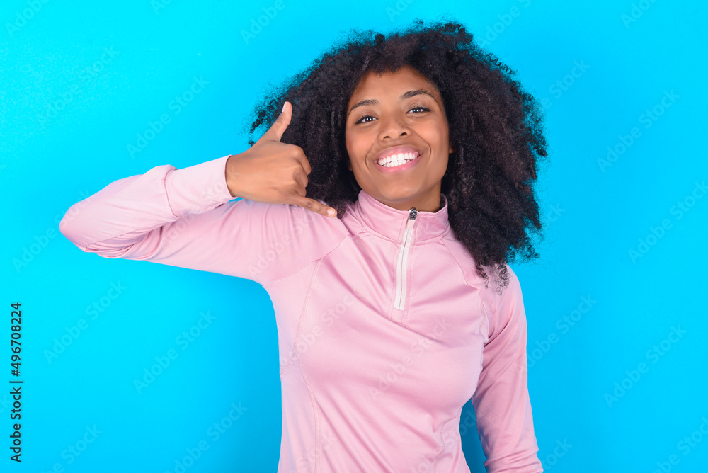 young woman with afro hairstyle in technical sports shirt against blue background makes phone gesture, says call me back again, has glad expression.