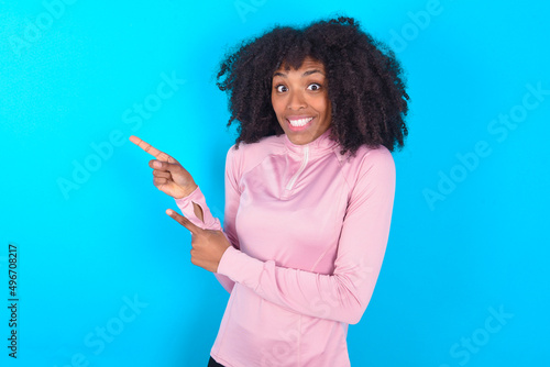 young woman with afro hairstyle in technical sports shirt against blue background points at copy space indicates for advertising gives right direction
