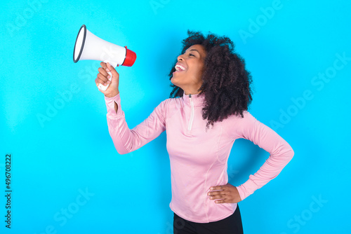 Funny young woman with afro hairstyle in technical sports shirt against blue background People sincere emotions lifestyle concept. Mock up copy space. Screaming in megaphone.