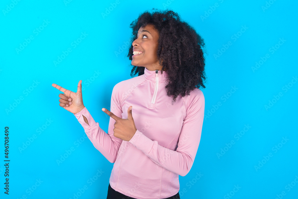 Optimistic young woman with afro hairstyle in technical sports shirt against blue background points with both hands and looking at empty space. Optimistic young woman with afro hairstyle in technical sports shirt against blue background points with both hands and looking at empty space.