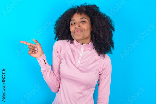 young woman with afro hairstyle in technical sports shirt against blue background points to side on blank space demonstrates advertisement. People and promotion concept