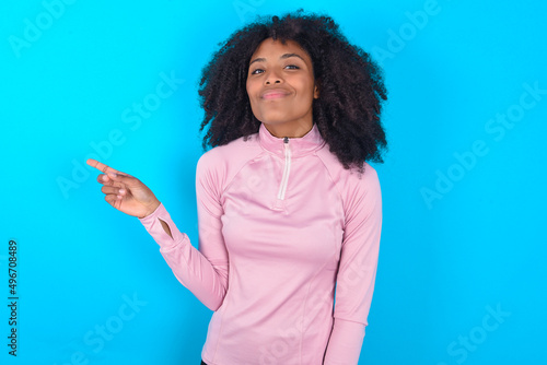 Positive young woman with afro hairstyle in technical sports shirt against blue background with satisfied expression indicates at upper right corner shows good offer suggests to click on link © Jihan
