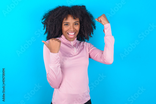 Hooray cool young woman with afro hairstyle in technical sports shirt against blue background point back empty space hand fist