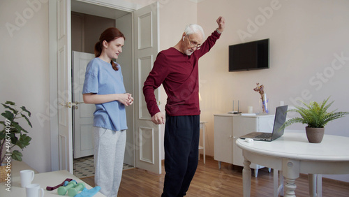 A physiotherapist watches as an old man swings his arms. He repeats the exercises that he sees on the laptop screen