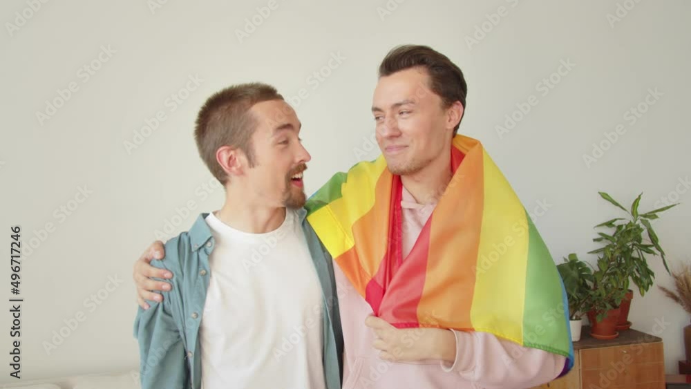 Two young men stand together, covered with a lgbt flag, and talk to each other, smiling Two young men stand together, covered with a lgbt flag, and talk to each other, smiling