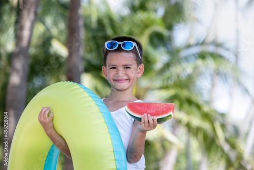 A boy wearing sunglasses holding a watermelon and a rubber ring smiles happily on his vacation.