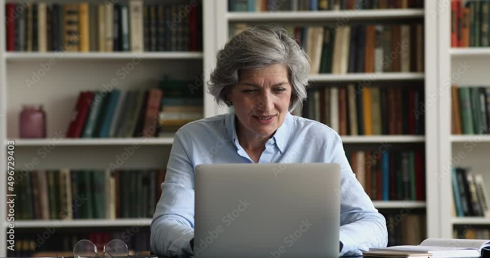 Busy smiling middle-aged woman sit at desk in cozy office or library work on laptop, make online research, prepare report, doing telecommute task feels motivated. Workflow using modern tech concept Busy smiling middle-aged woman sit at desk in cozy office or library work on laptop, make online research, prepare report, doing telecommute task feels motivated. Workflow using modern tech concept