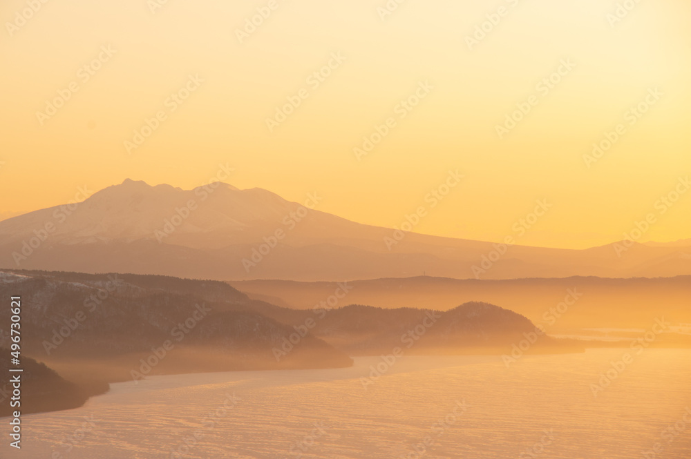 薄いオレンジ色の夜明けの空の下の霞んだ山々のシルエットと湖の風景。