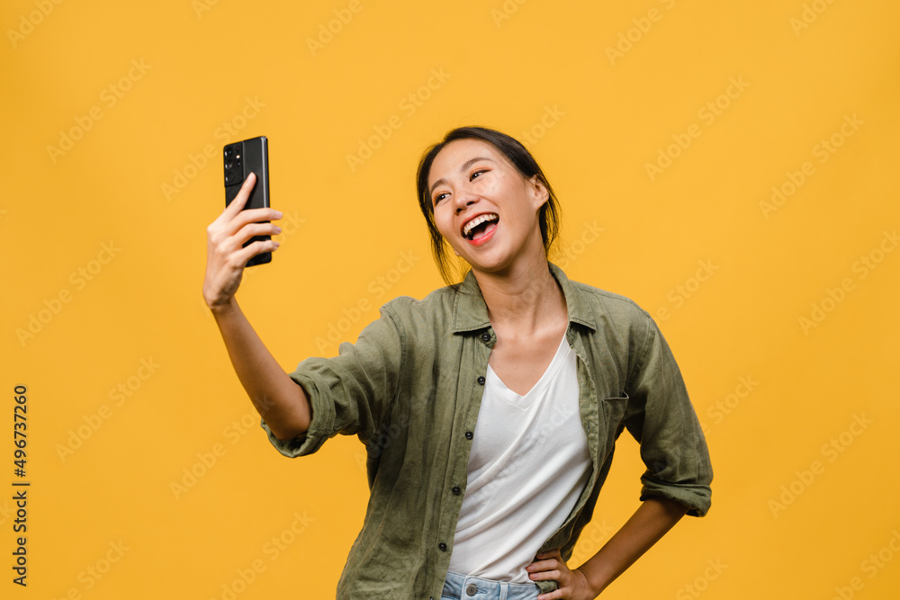 Smiling adorable Asian female making selfie photo on smart phone with positive expression in casual clothing and stand isolated on yellow background. Happy adorable glad woman rejoices success. Smiling adorable Asian female making selfie photo on smart phone with positive expression in casual clothing and stand isolated on yellow background. Happy adorable glad woman rejoices success.