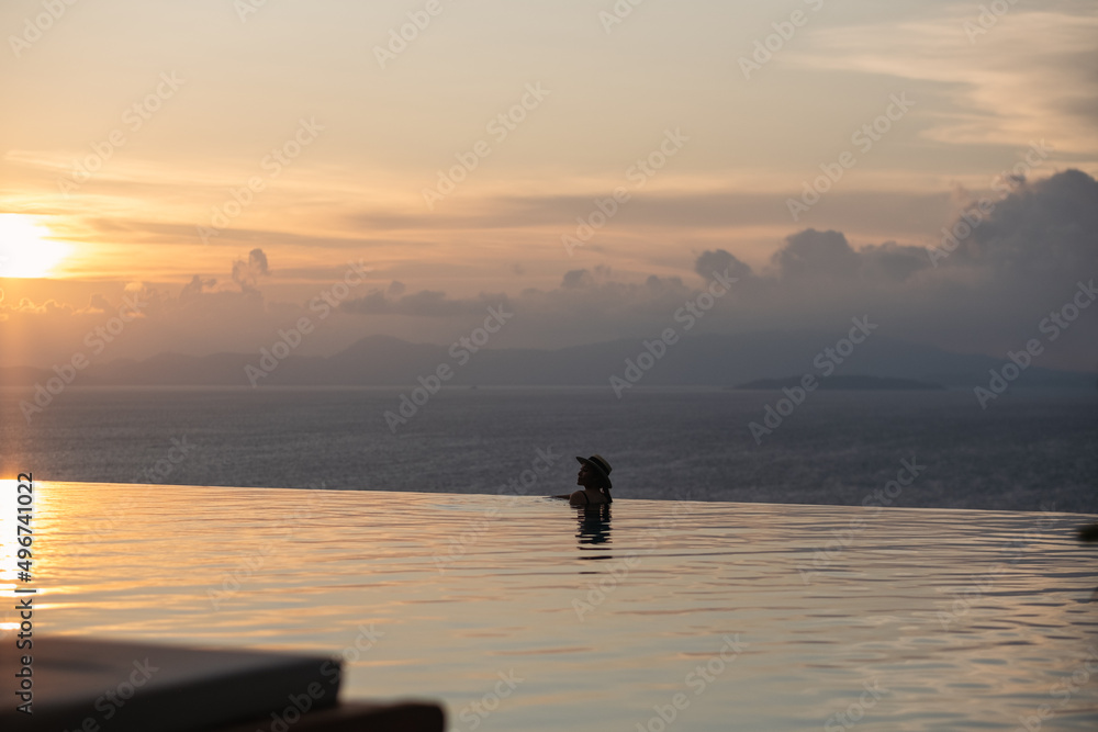 A woman with hat relaxing in infinity swimming pool and looking at a beautiful sunset and the sea view