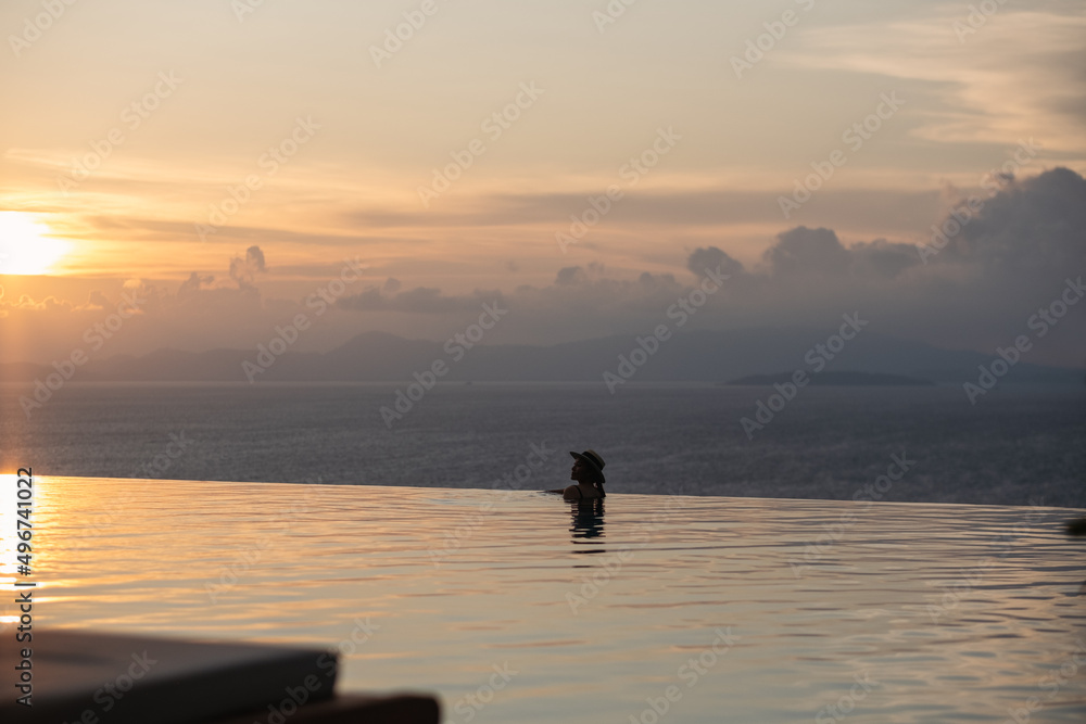 A woman with hat relaxing in infinity swimming pool and looking at a beautiful sunset and the sea view A woman with hat relaxing in infinity swimming pool and looking at a beautiful sunset and the sea view
