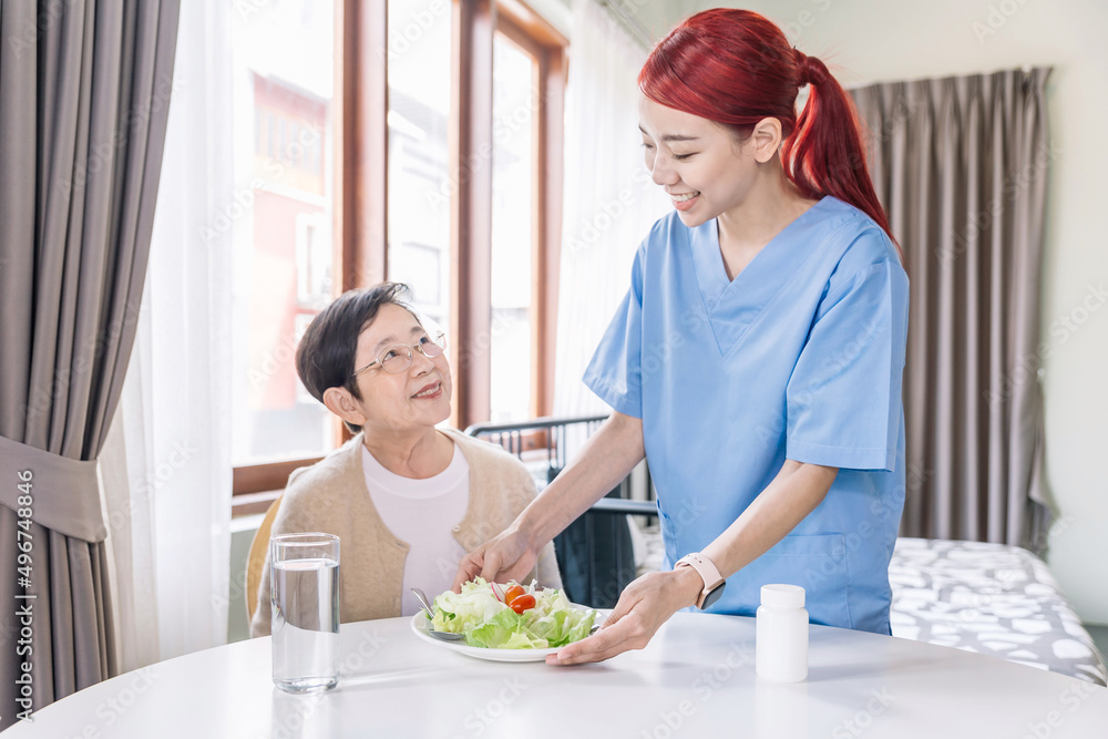 Assistant Asian woman caregiver giving care aging woman adult by serving a salad eating meals at home. Caregiver visit at home. Home health care and nursing home concept.