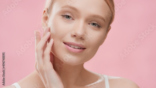 Young slim good-looking European woman with light hair in white bikini turns her head to the camera touching her jawline and smiling wide against pink ripple background | Skin moisturizing promotion