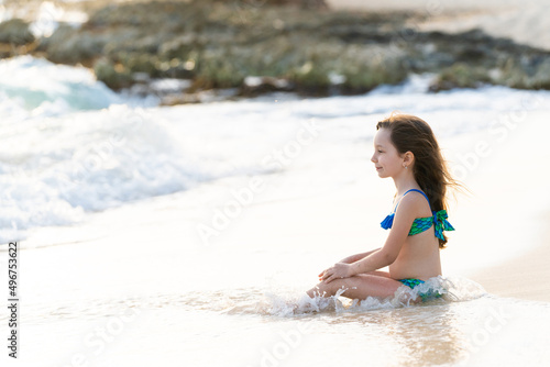 A little girl 6 years old in a mermaid swimsuit sits on a sea rocky coast. The wind blows hair and waves