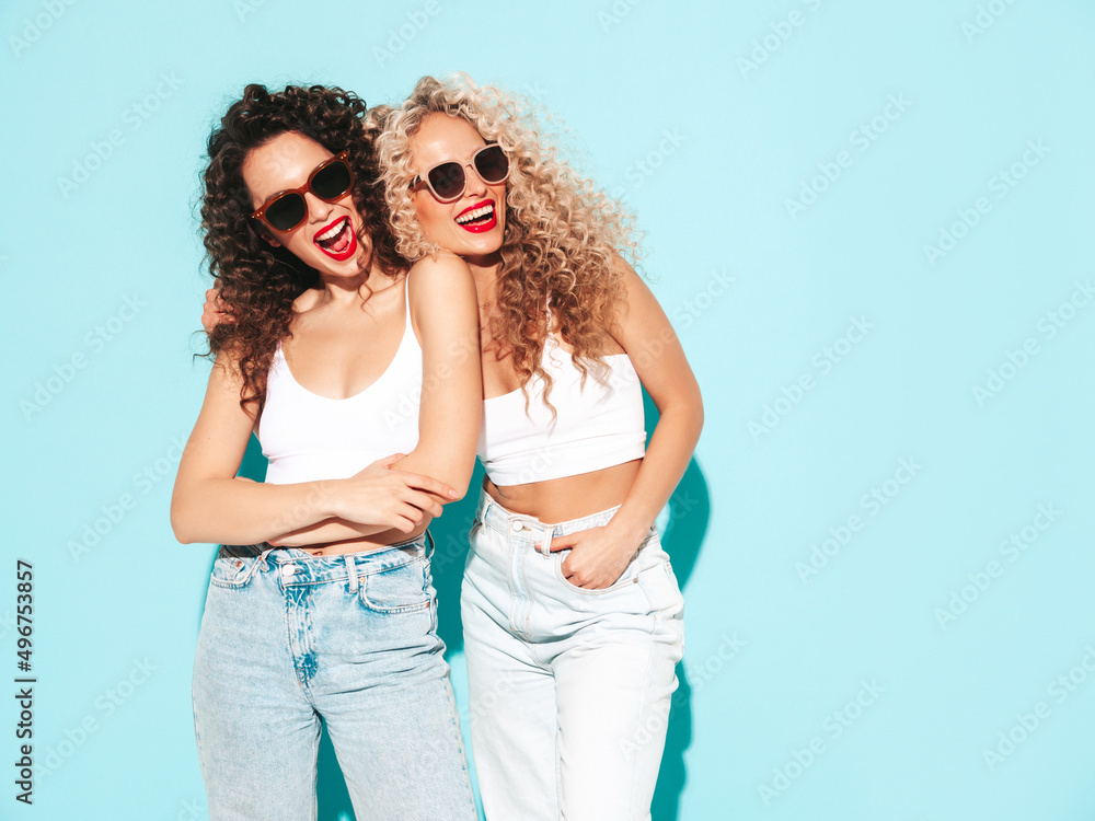 Two beautiful sexy smiling hipster women in summer clothes. Trendy models with afro curls hairstyle having fun in studio. Hot female isolated on blue. Happy and cheerful. In sunglasses Two beautiful sexy smiling hipster women in summer clothes. Trendy models with afro curls hairstyle having fun in studio. Hot female isolated on blue. Happy and cheerful. In sunglasses