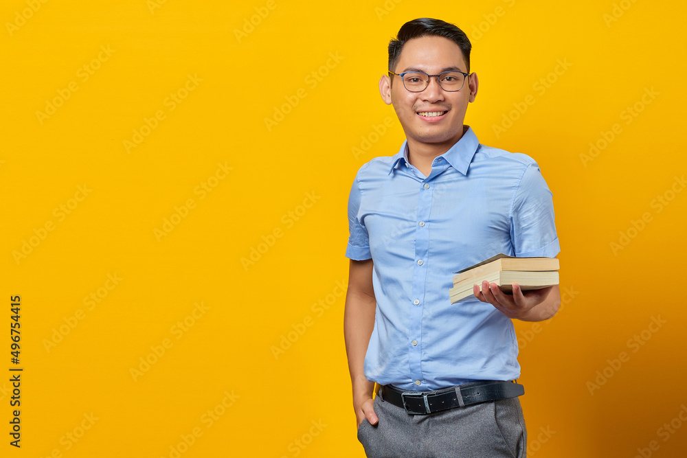 Fototapeta premium portrait of smiling young asian man in glasses holding book isolated on yellow background. businessman and entrepreneur concept