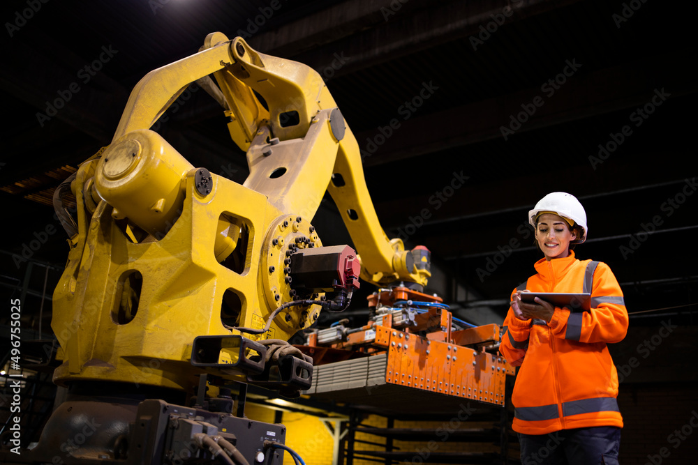 Fototapeta premium Female factory worker in safety equipment standing by industrial robot machine and watching manufacturing process on tablet computer.
