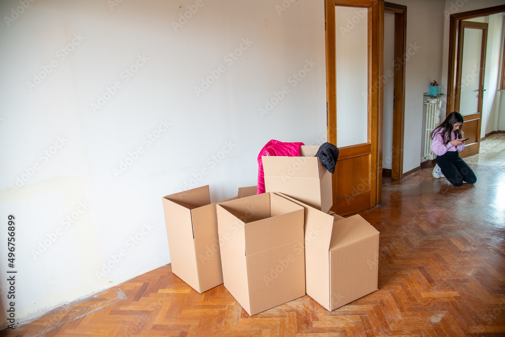 Moving to a new home concept. Cardboard boxes along the parquet floor of a empty room with a young girl seated on the background. Moving to a new home concept. Cardboard boxes along the parquet floor of a empty room with a young girl seated on the background.