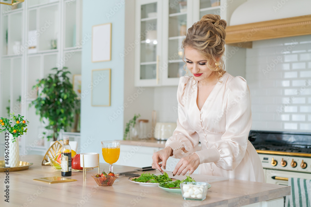 Happy luxurious woman prepares light salad with strawberries and arugula with cheese against backdrop bright kitchen at home in beautiful delicate dress with makeup and hair. Festive mood in morning
