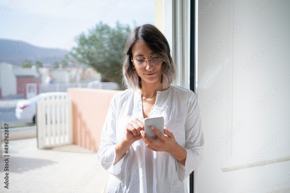 Young woman wearing eyeglasses surfing net through smart phone standing at doorway Young woman wearing eyeglasses surfing net through smart phone standing at doorway
