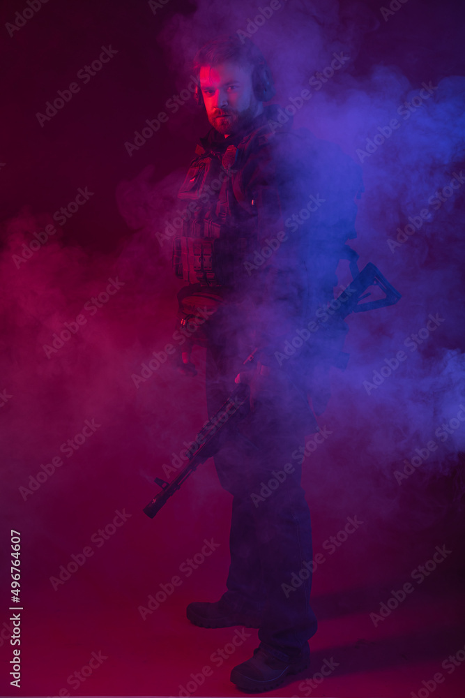 soldier in full gear with weapons. a man in headphones, body armor, with a backpack and a belt. red background. colored, blue-red light. smoke around the military. explosion, chemical attack