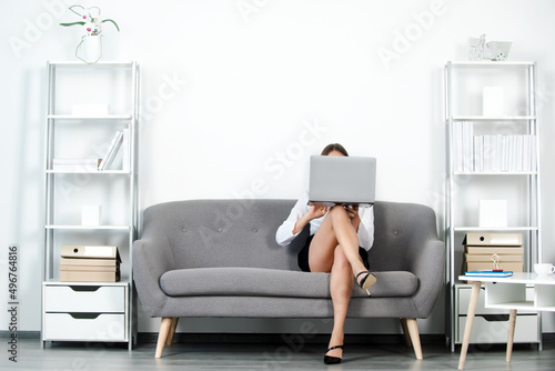 Beautiful secretary legs. Seductive sexy secretary freelancer cover face with laptop computer and sexy legs sit on sofa in office. Sexy business woman. Sensual young girl employee.