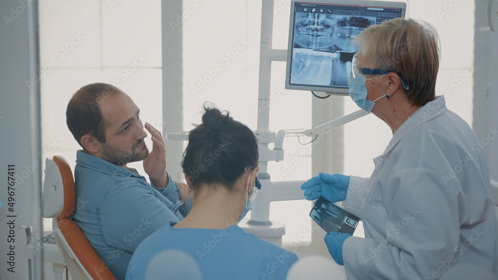 Obraz premium Dentist and nurse explaining x ray scan to patient with toothache in stomatology cabinet. Team of specialists doing oral care consultation to treat man in pain with denture problem.