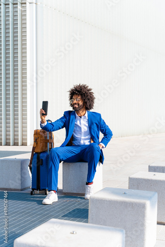 Smiling businessman with luggage taking selfie through smart phone sitting on concrete block photo