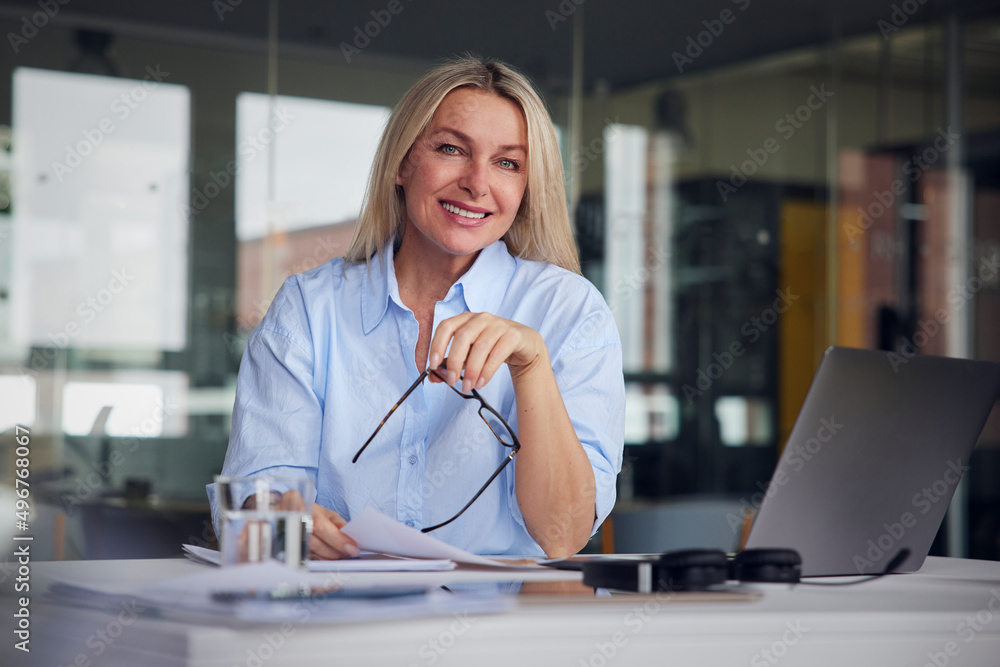 Smiling businesswoman holding paper and eyeglasses sitting by laptop at desk in office Smiling businesswoman holding paper and eyeglasses sitting by laptop at desk in office