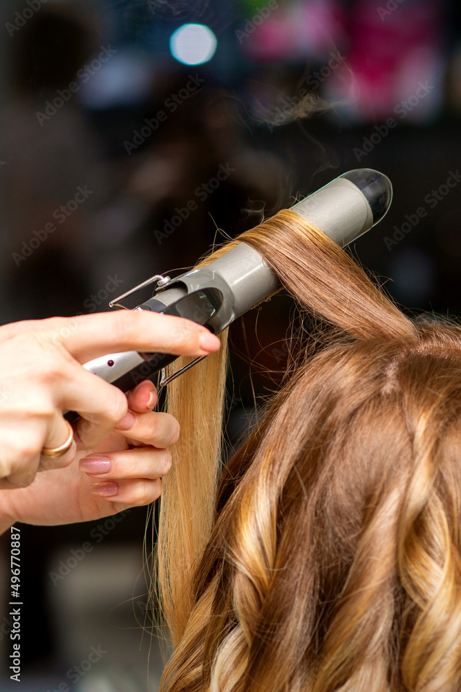Hairdresser makes curls with a curling iron for the young woman with long brown hair in a beauty salon Hairdresser makes curls with a curling iron for the young woman with long brown hair in a beauty salon
